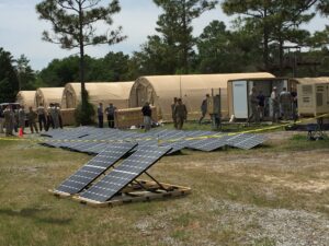 Military personnel near portable solar panels and tents in a wooded area.